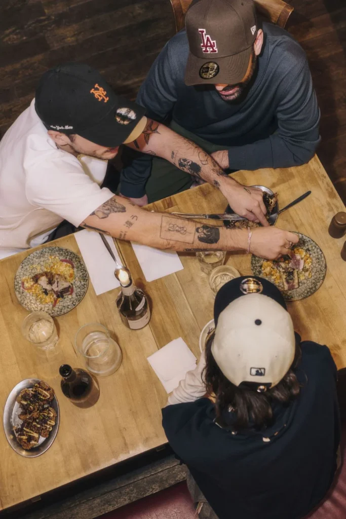 Três amigos a jantar, usando bonés de baseball, à volta da mesa.