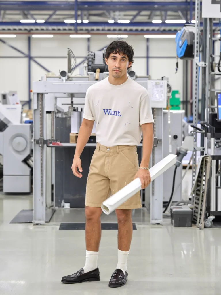 Homem em fábrica com calções, t-shirt e projeto enrolado.