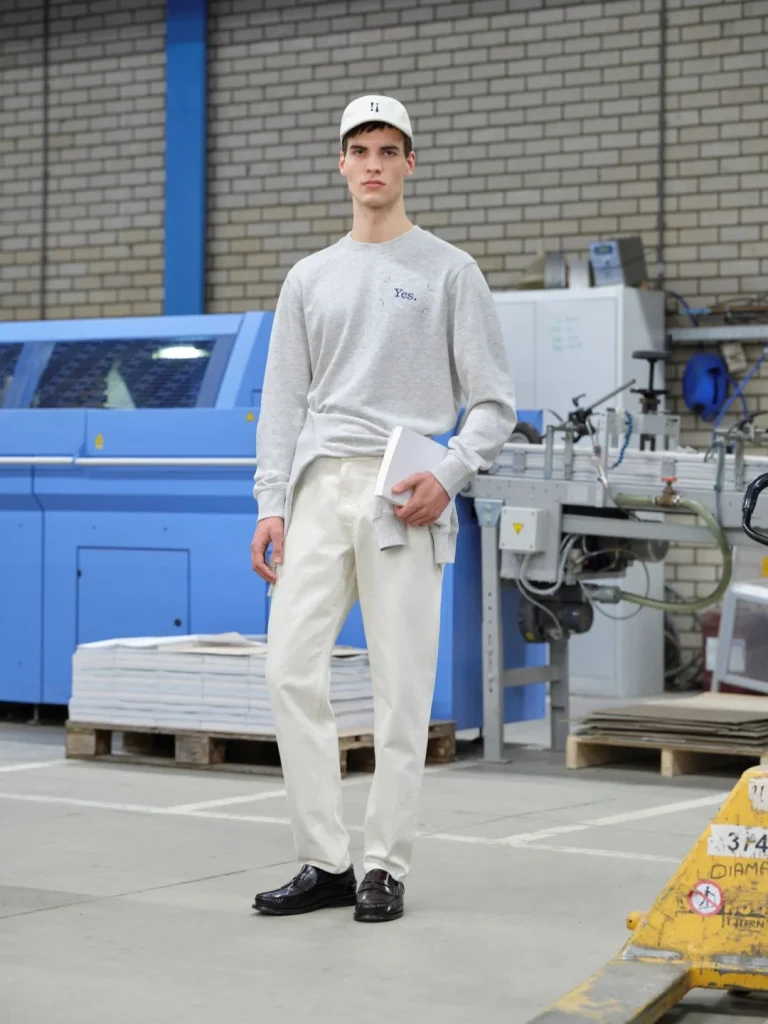 Homem com boné e sweatshirt em fábrica, segurando livro.