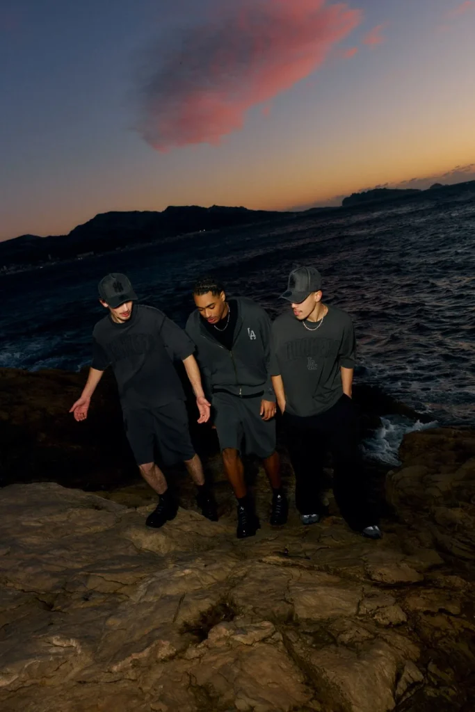 Três homens com roupa escura junto ao mar ao entardecer.