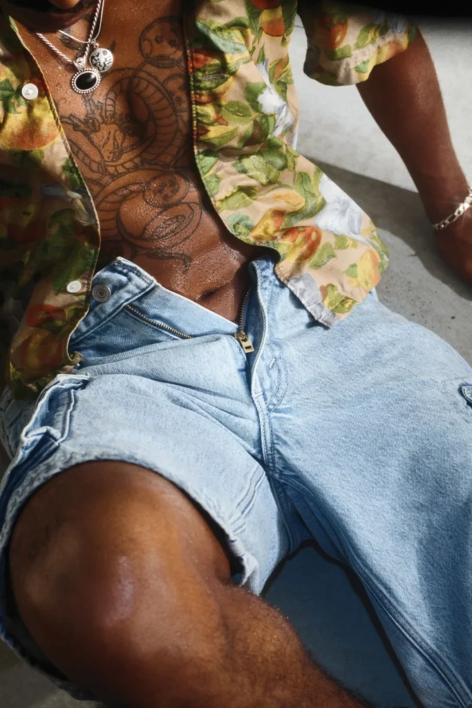 Torso de pessoa com tatuagens, camisa floral aberta e calças de ganga.