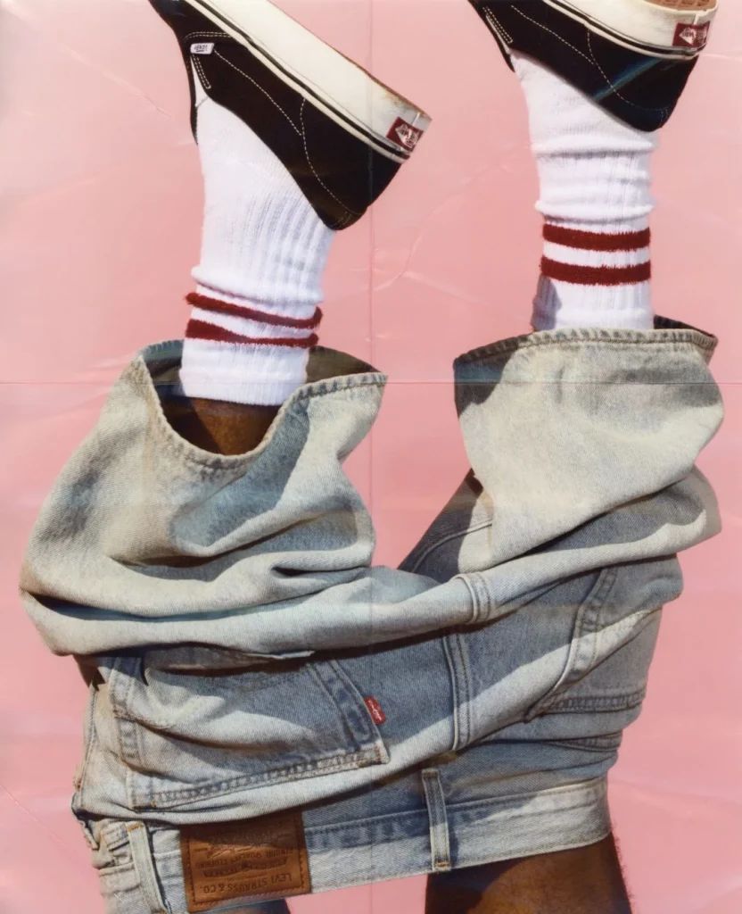 Pés com Vans, meias listradas e jeans Levi's contra fundo rosa.