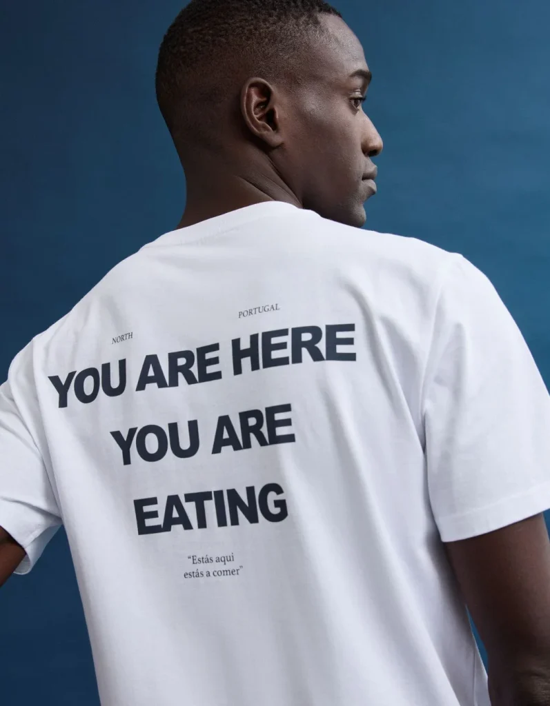Homem de costas com t-shirt branca e texto 'YOU ARE HERE YOU ARE EATING'.