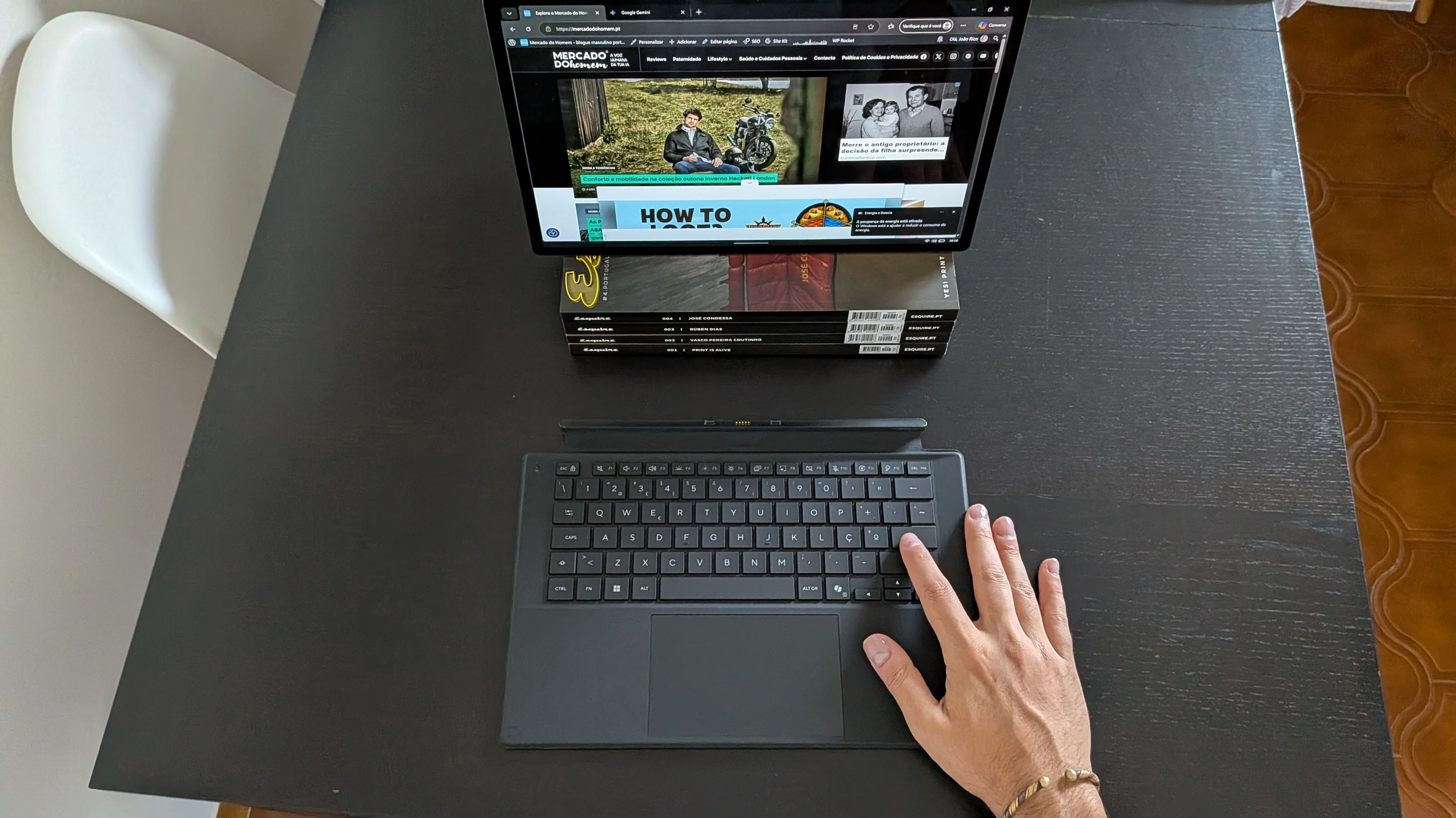 Ecrã do tablet Asus ProArt PZ14 a exibir o blogue apoiado em revistas com o teclado destacado na mesa e uma mão pousada nas teclas.