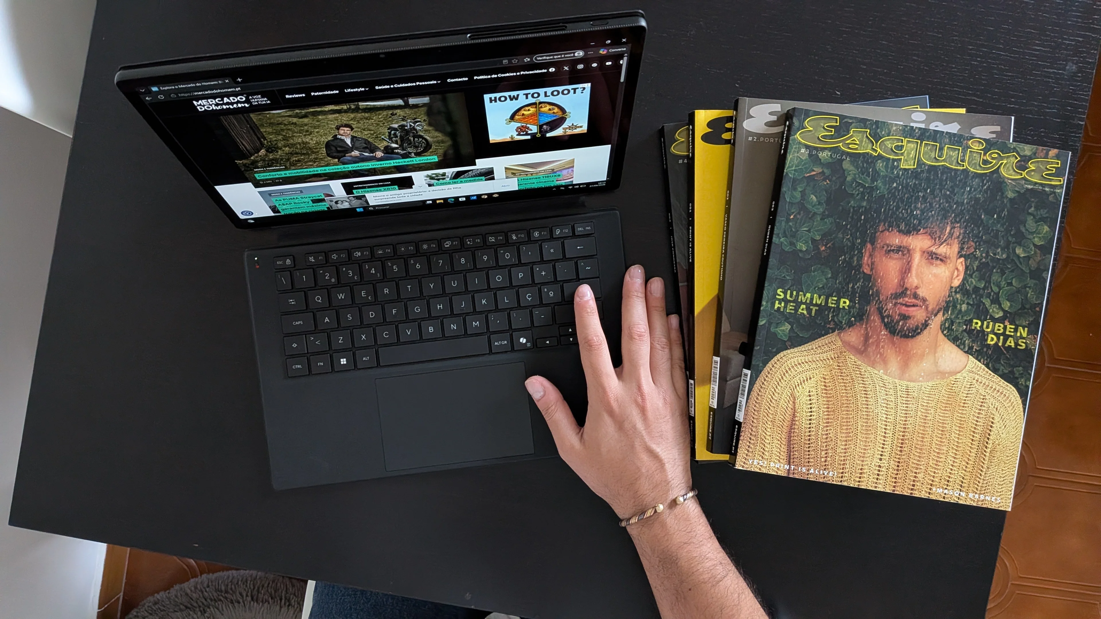 Mão sobre o teclado do Asus ProArt PZ14 com o site Mercado do Homem no ecrã ao lado de revistas Esquire numa mesa preta.