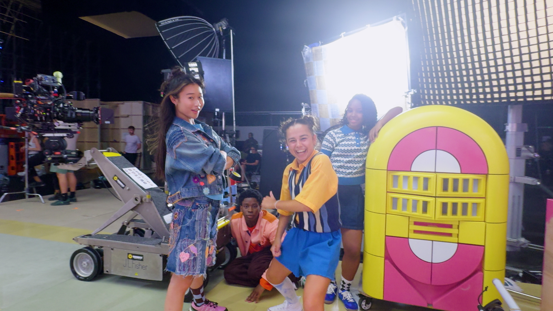 Quatro jovens atores nos bastidores do filme live action LEGO Friends junto a uma jukebox colorida e equipamentos de câmara
