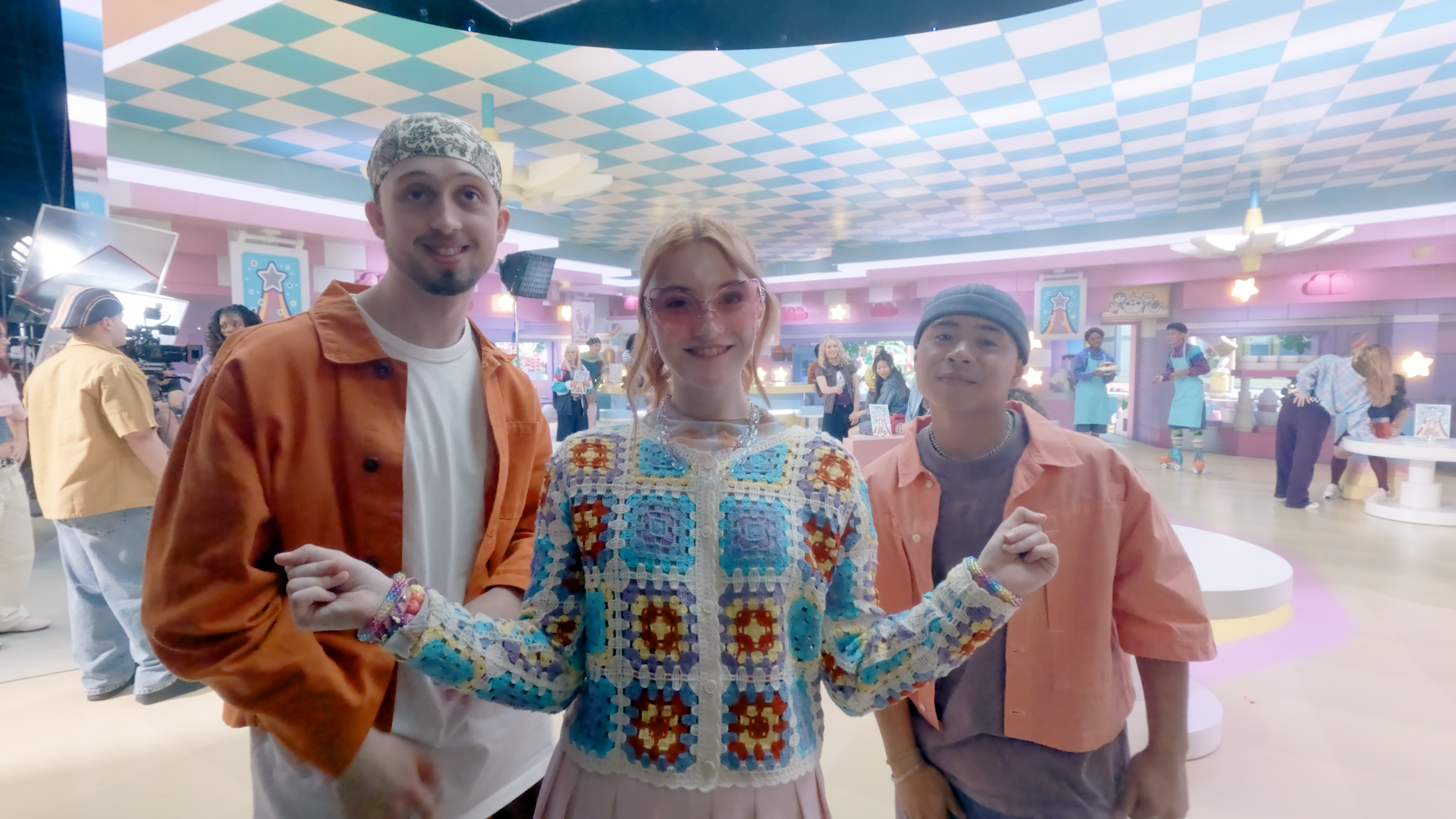 Três jovens atores sorridentes no cenário de bastidores do filme live action lego friends. A rapariga ao centro veste um casaco de croché colorido e óculos cor-de-rosa, acompanhada por dois rapazes.