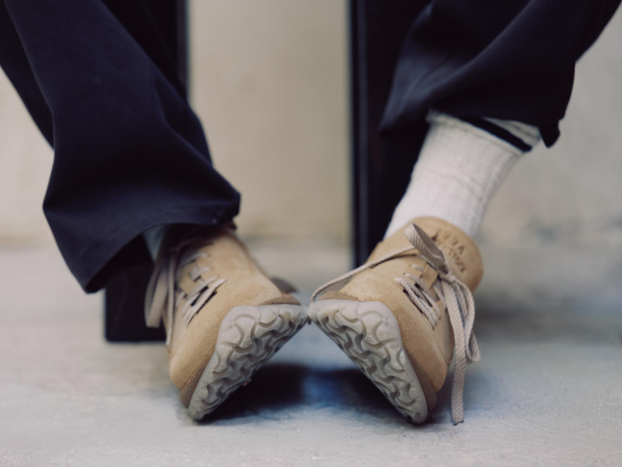 Pés a usar o modelo híbrido Birkenstock 1774 Uerzell em camurça bege com atacadores, meias brancas e calças escuras, mostrando a sola texturizada.