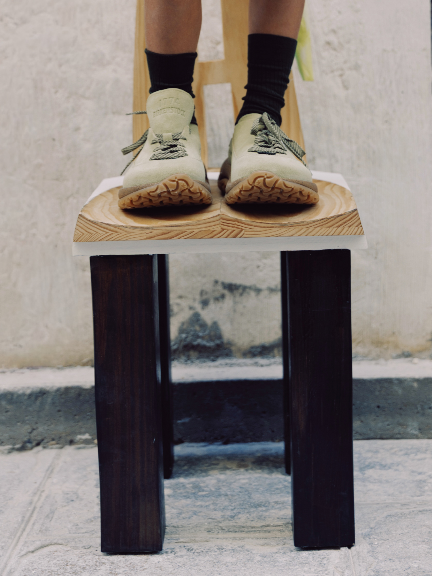 Pessoa em pé sobre uma cadeira de madeira a calçar o modelo Birkenstock 1774 Uerzell em camurça clara com meias pretas.