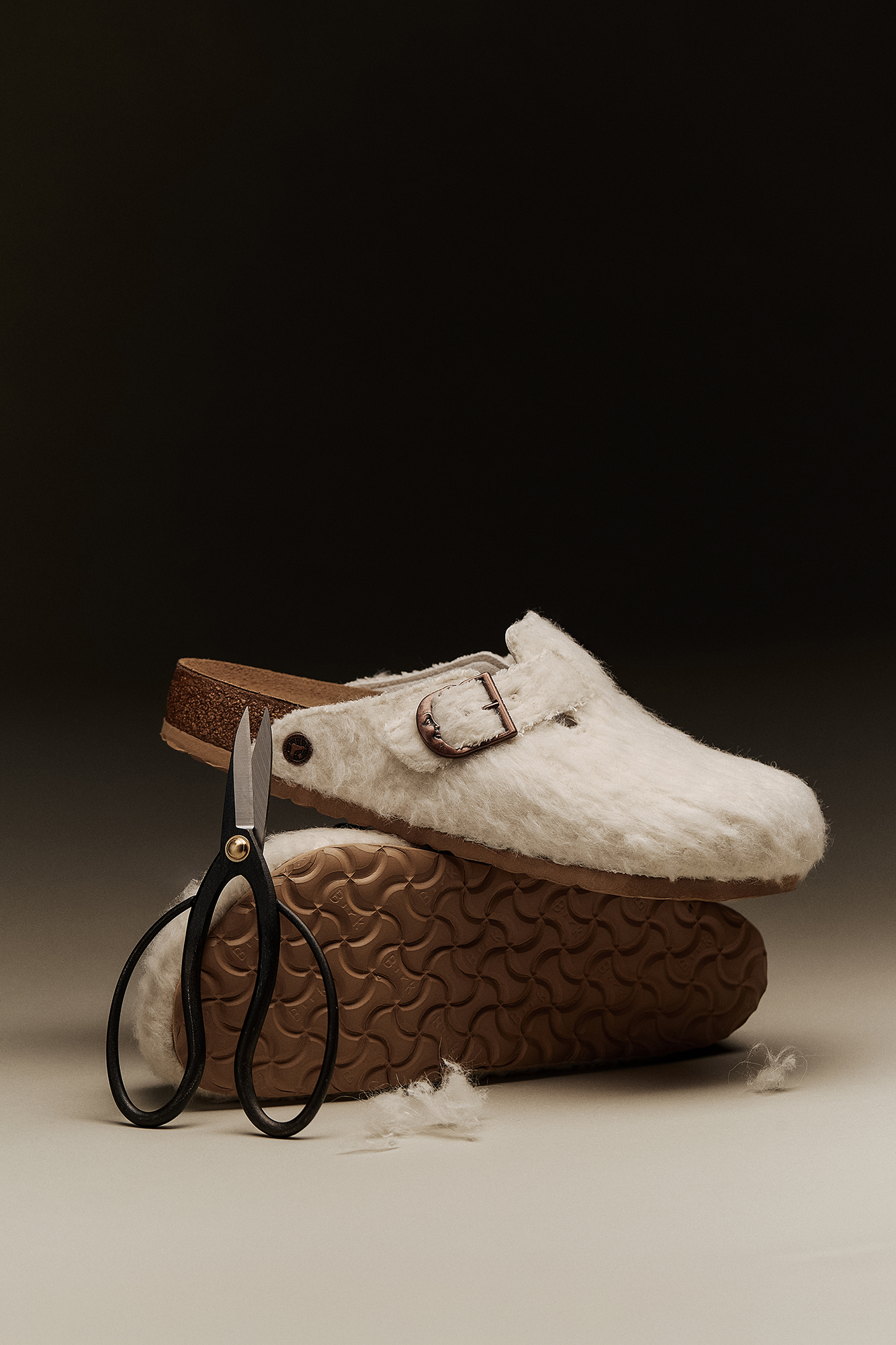 Par de socas Birkenstock Boston Cole Bennett em feltro off-white com fivela de lua crescente em cobre, empilhadas ao lado de uma tesoura preta e restos de lã.