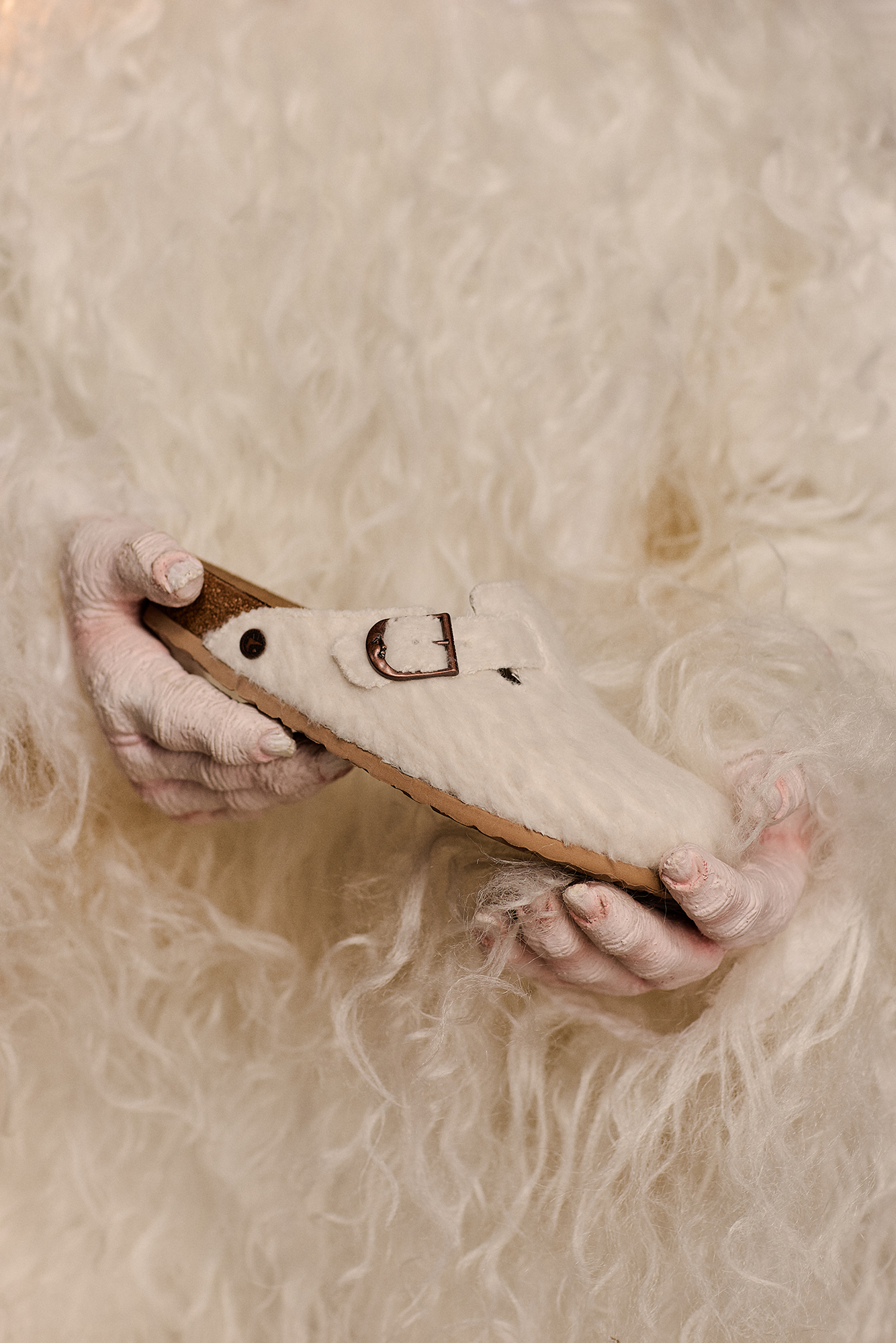 Mãos pintadas de branco seguram o tamanco Birkenstock Boston Cole Bennett em feltro off-white contra um fundo de lã felpuda.