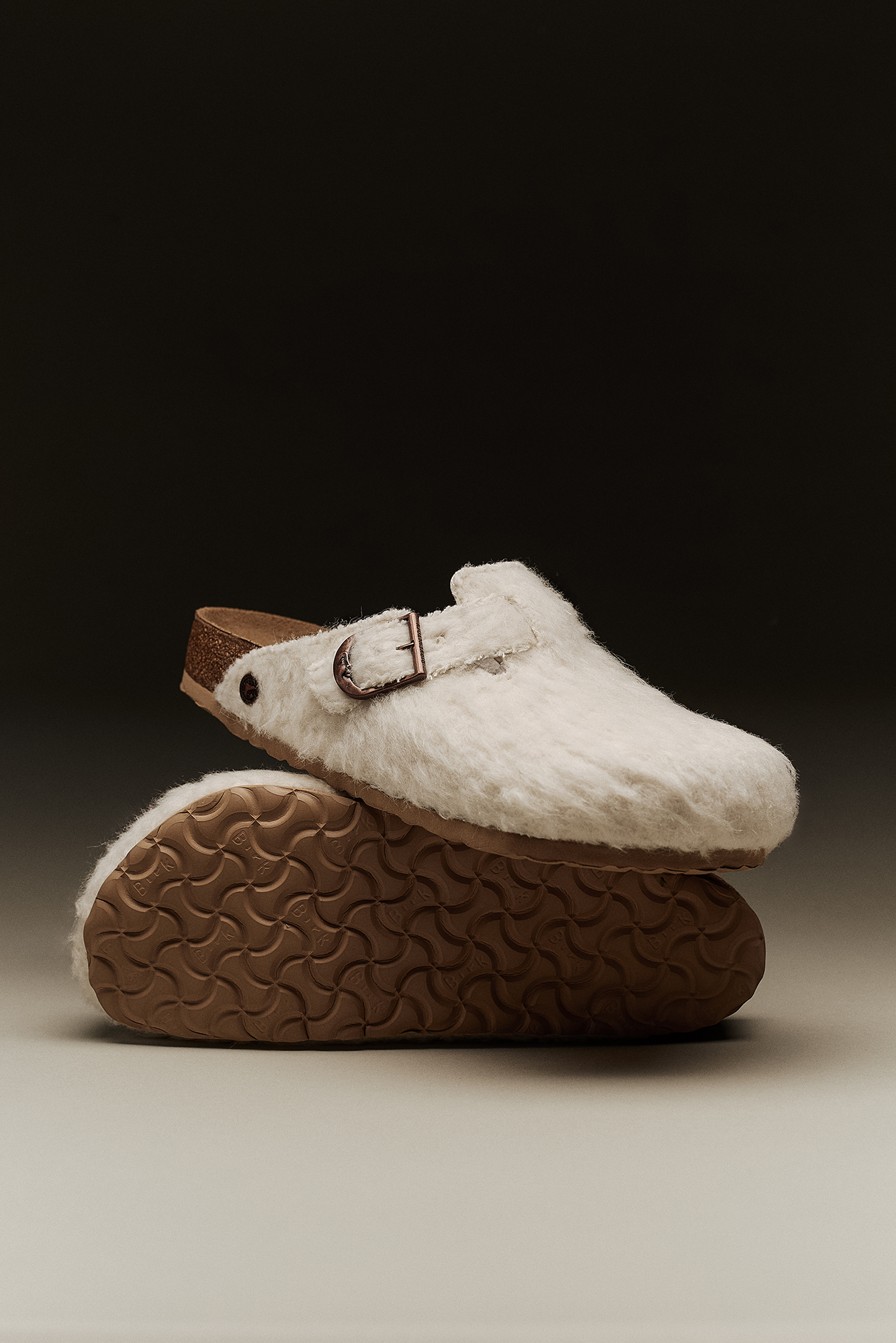 Par de socos Birkenstock Boston Cole Bennett em feltro de lã off-white empilhados, destacando a textura felpuda, a sola castanha e a fivela em cobre.