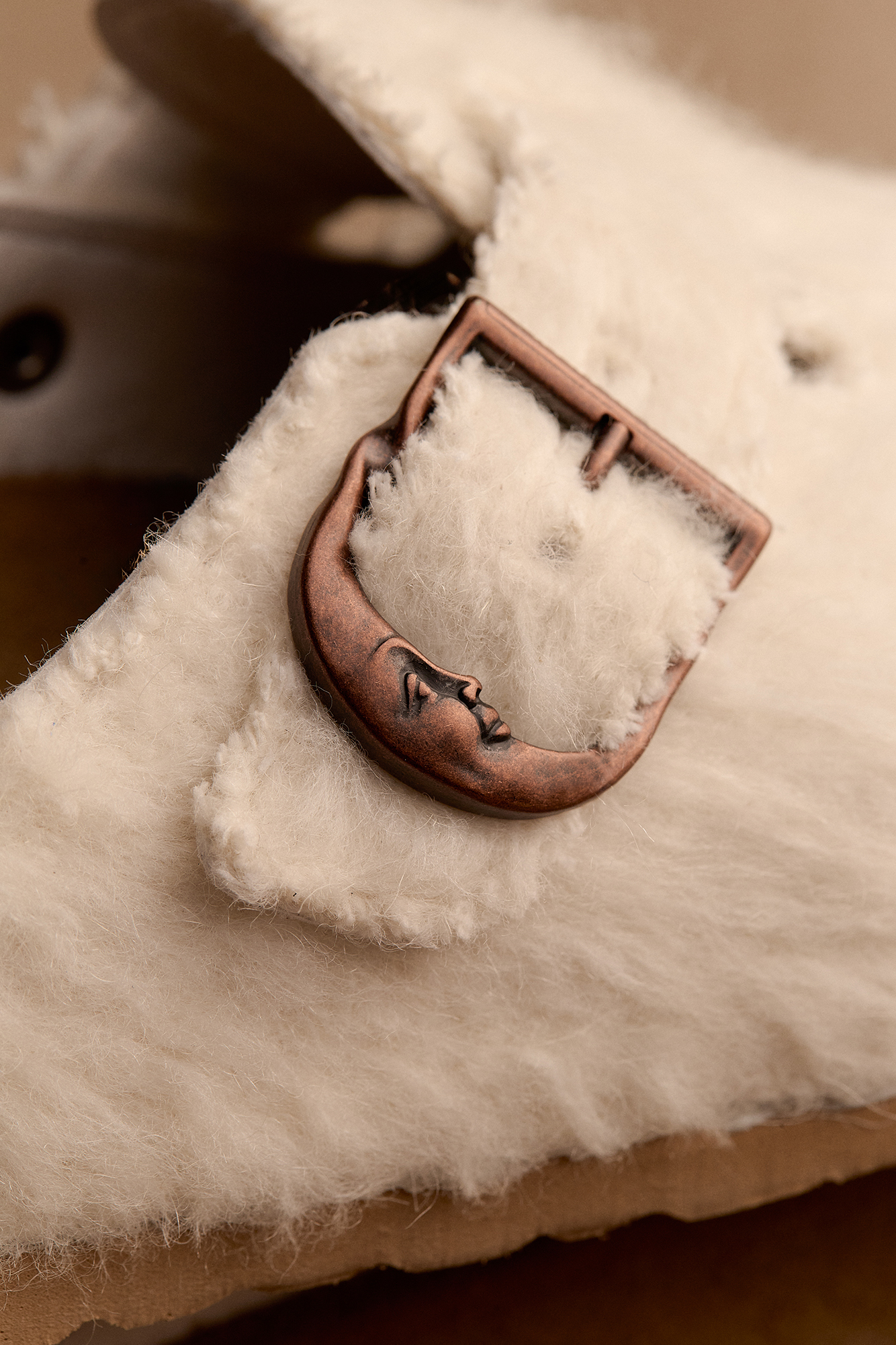 Detalhe aproximado da fivela em forma de lua crescente com rosto em cobre envelhecido sobre o feltro de lã off-white do modelo Birkenstock Boston Cole Bennett