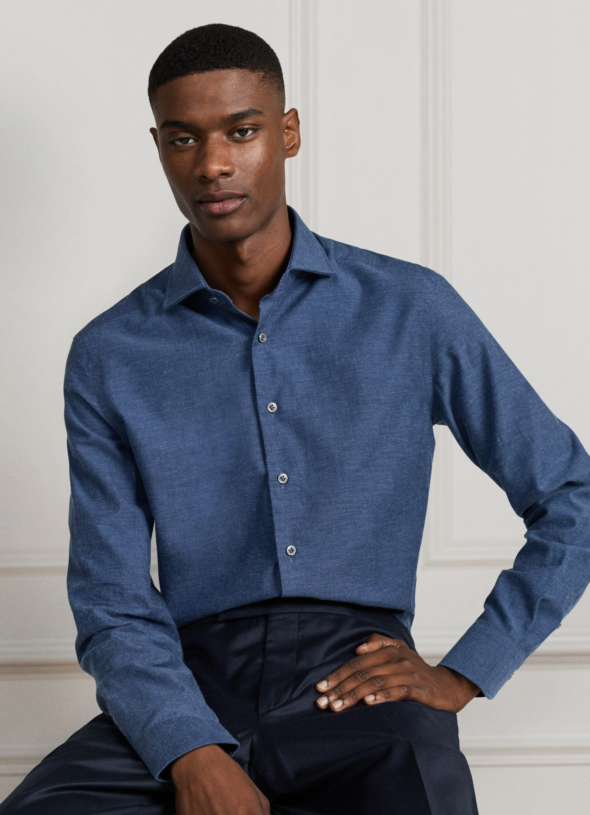 Homem sentado a vestir uma camisa azul marinho texturizada e calças escuras da coleção de inverno 2026.