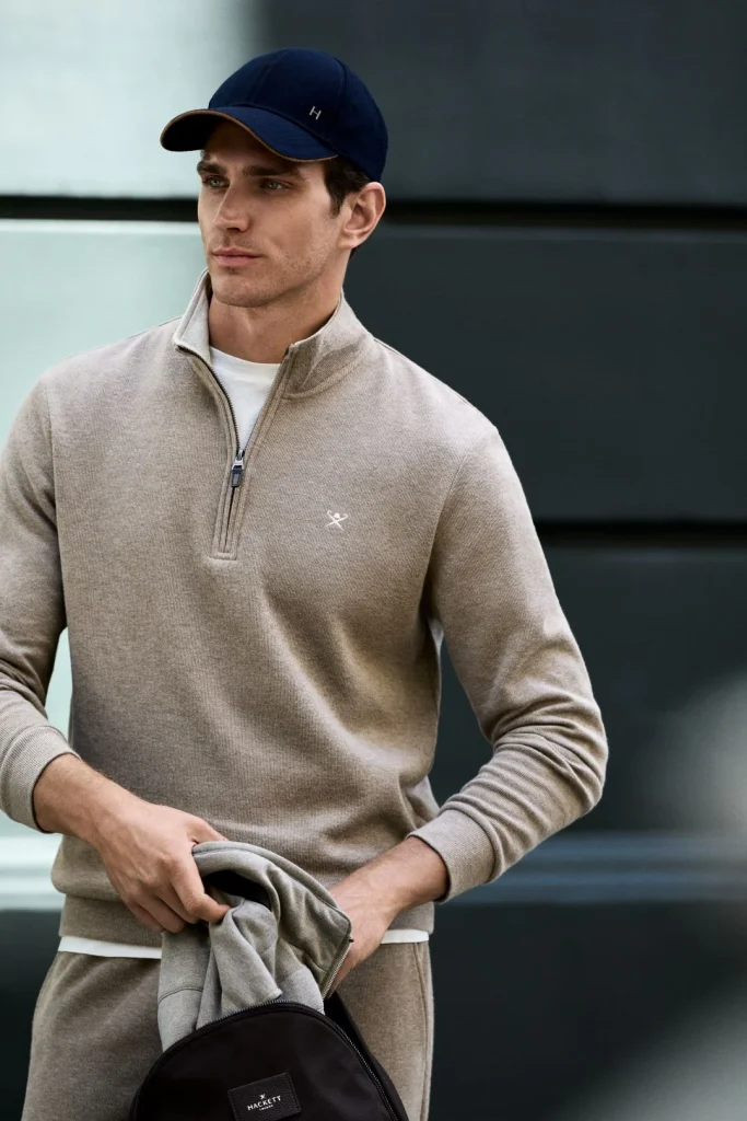 Homem veste camisola bege de meio fecho e boné azul marinho da coleção Hackett London outono inverno num ambiente urbano.
