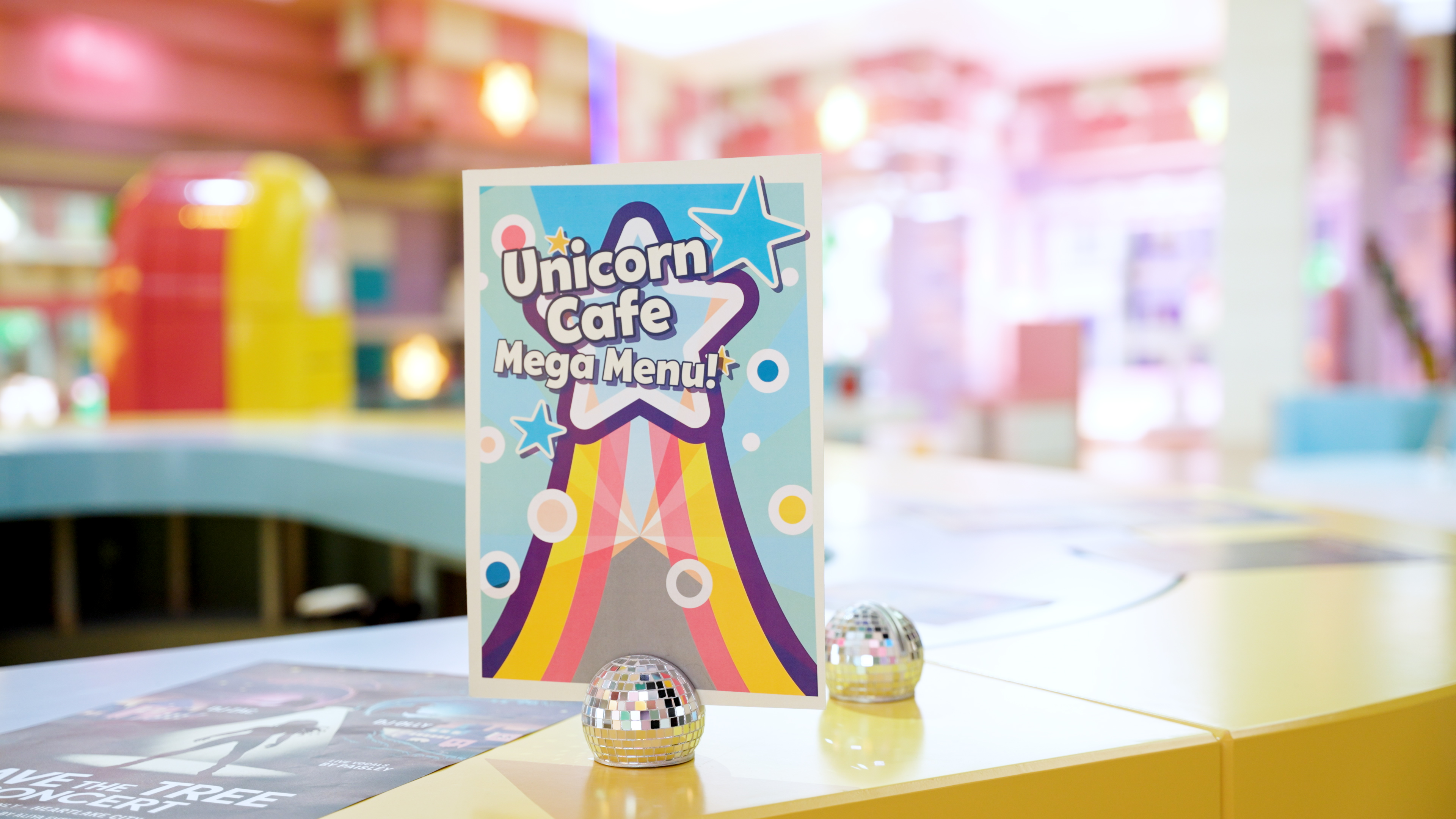 Cartaz com o texto Unicorn Cafe Mega Menu apoiado em pequenas bolas de espelhos num balcão amarelo no cenário do filme live action LEGO Friends