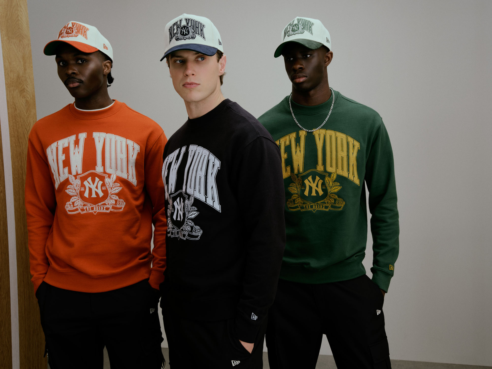 Três homens vestem sweatshirts oversized e bonés 9FORTY da coleção New Era MLB New York Yankees Clubhouse em laranja, azul-marinho e verde