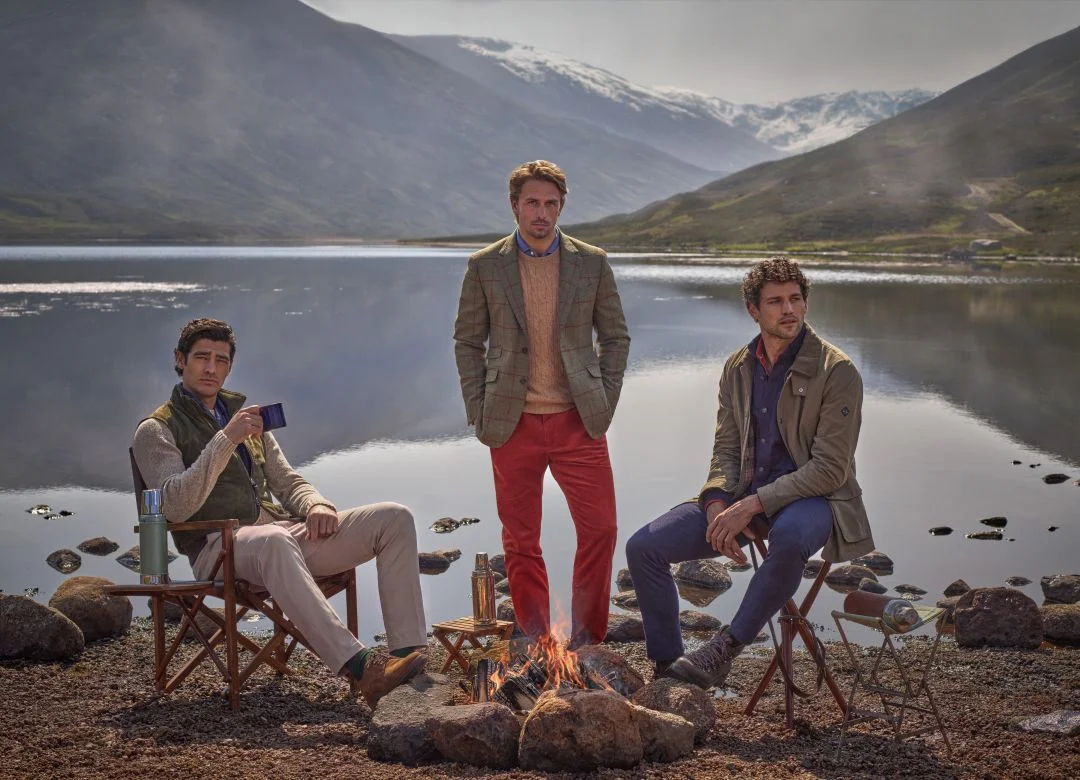 Três homens vestem casacos de tweed e malhas da coleção outono inverno Hackett London enquanto relaxam junto a uma fogueira na margem de um lago nas Terras Altas da Escócia.