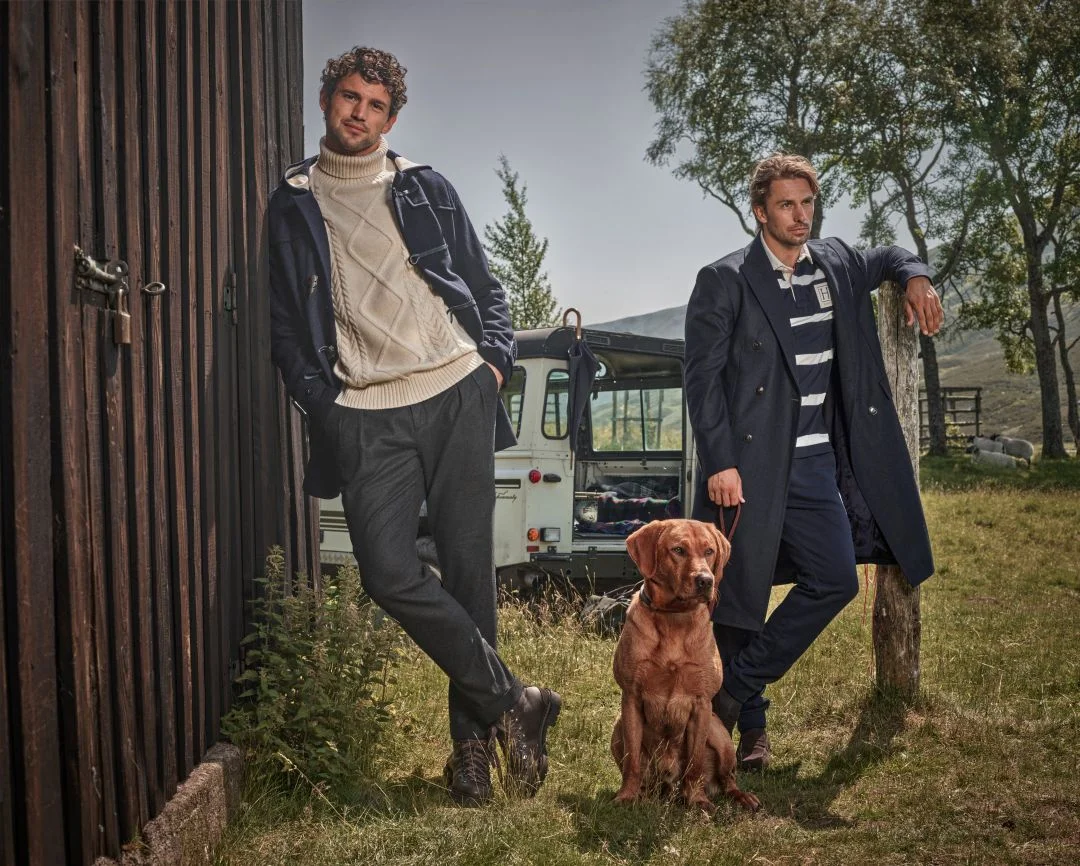 Dois homens vestem malhas e casacos da coleção outono inverno Hackett London num campo com um cão labrador e um jipe clássico ao fundo.