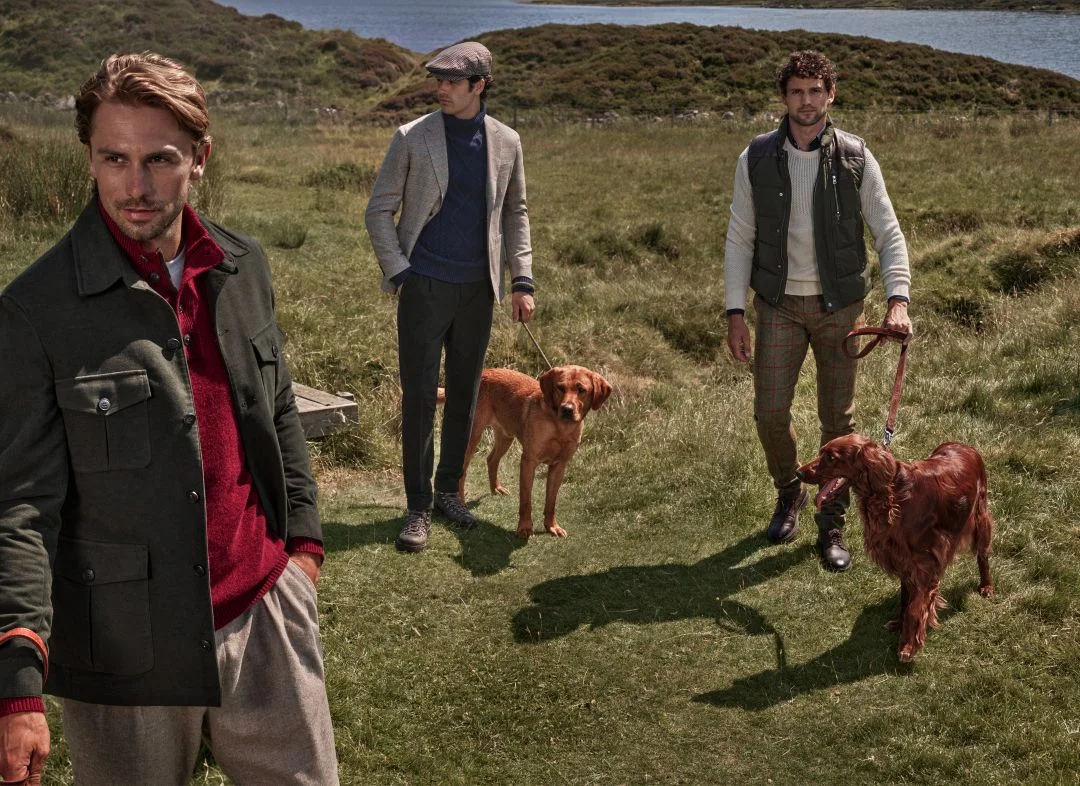 Três homens com cães numa paisagem rural escocesa a vestir peças da coleção outono inverno Hackett London com casacos, malhas e calças de tweed