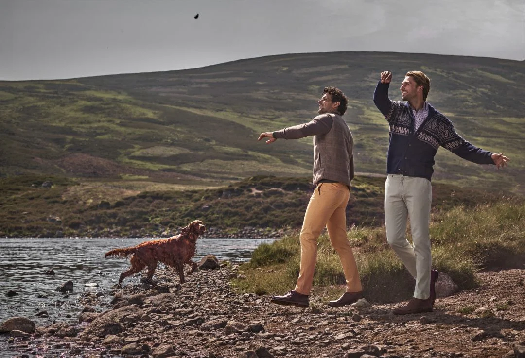 Dois homens a brincar com um cão junto a um lago nas Terras Altas da Escócia, a usar malhas e casacos da coleção outono inverno Hackett London.