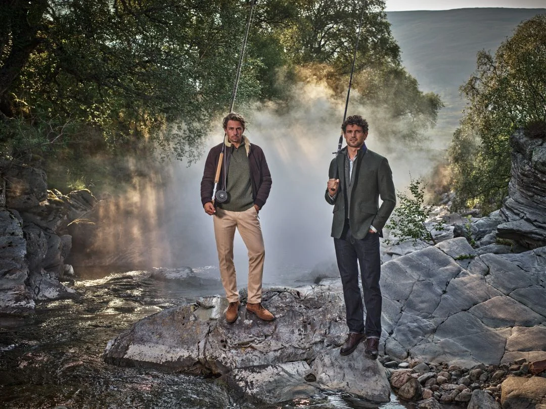 Dois homens com canas de pesca sobre rochas num rio enevoado vestem a coleção outono inverno Hackett London com blazer de tweed, casaco bomber e calças de ganga.