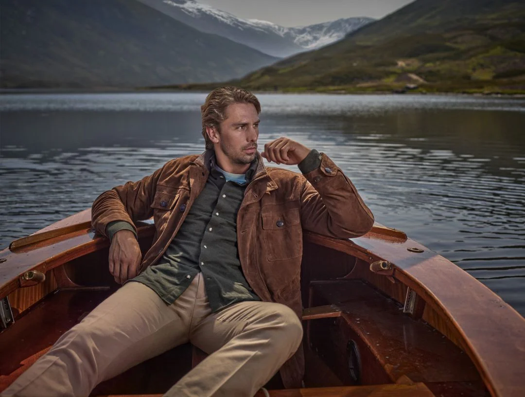 Homem sentado num barco de madeira num lago, a vestir um casaco castanho, camisa verde e calças beges da coleção outono inverno Hackett London, com montanhas ao fundo.