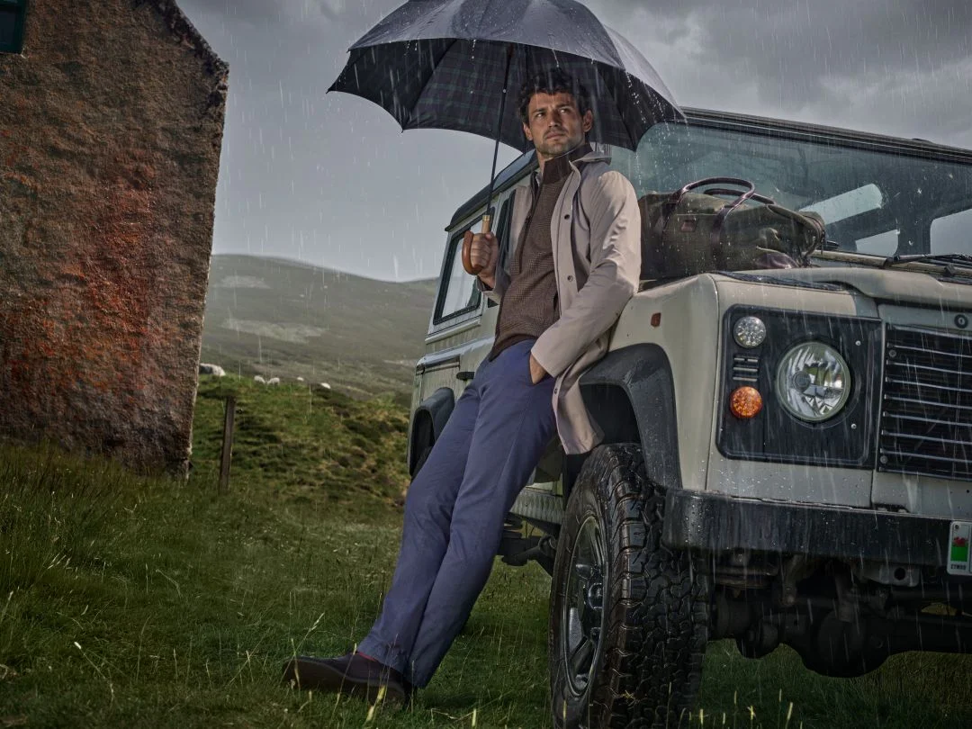 Homem com guarda-chuva encostado a um jipe sob chuva nas Terras Altas da Escócia, vestindo um sobretudo bege e camisola castanha da coleção outono inverno Hackett London.
