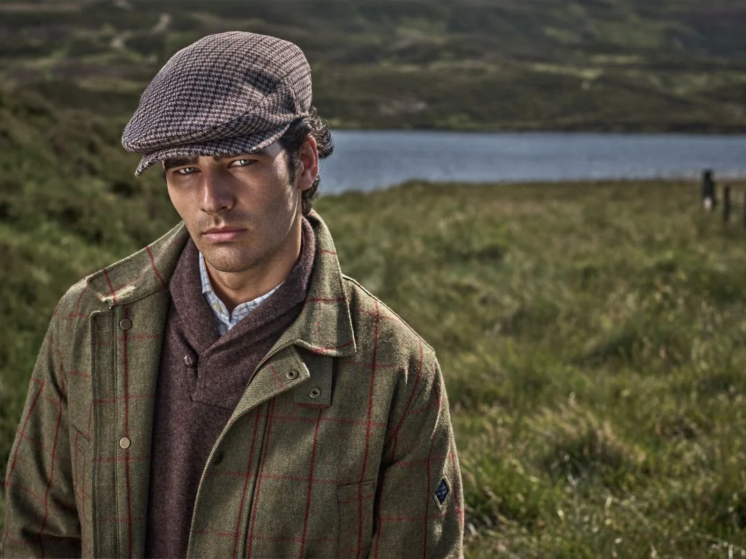 Homem com boina clássica, casaco verde com padrão xadrez e camisola de malha castanha numa paisagem rural escocesa, destacando a coleção outono inverno Hackett London.