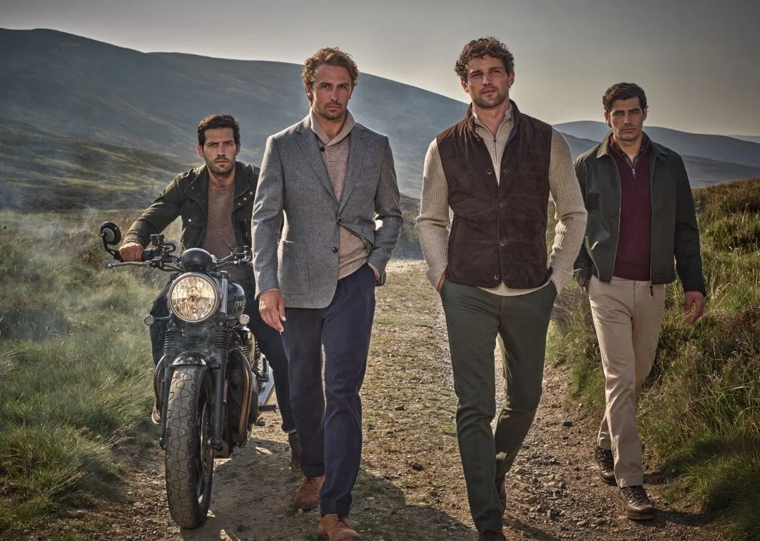 Quatro homens com roupas da coleção outono inverno Hackett London numa estrada de terra nas Terras Altas da Escócia ao lado de uma mota clássica.