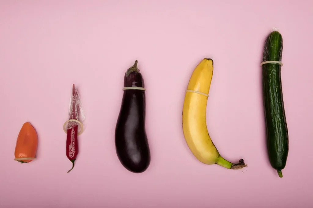 Cinco vegetais e frutas de diferentes espessuras com preservativos num fundo rosa representando a largura nominal.
