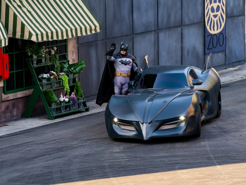 Batman a acenar ao lado do Batmobile numa rua.