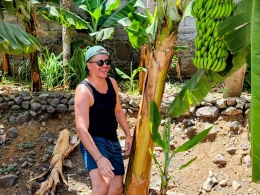 Homem a sorrir junto de uma bananeira com bananas verdes.