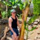 Homem a sorrir junto de uma bananeira com bananas verdes.