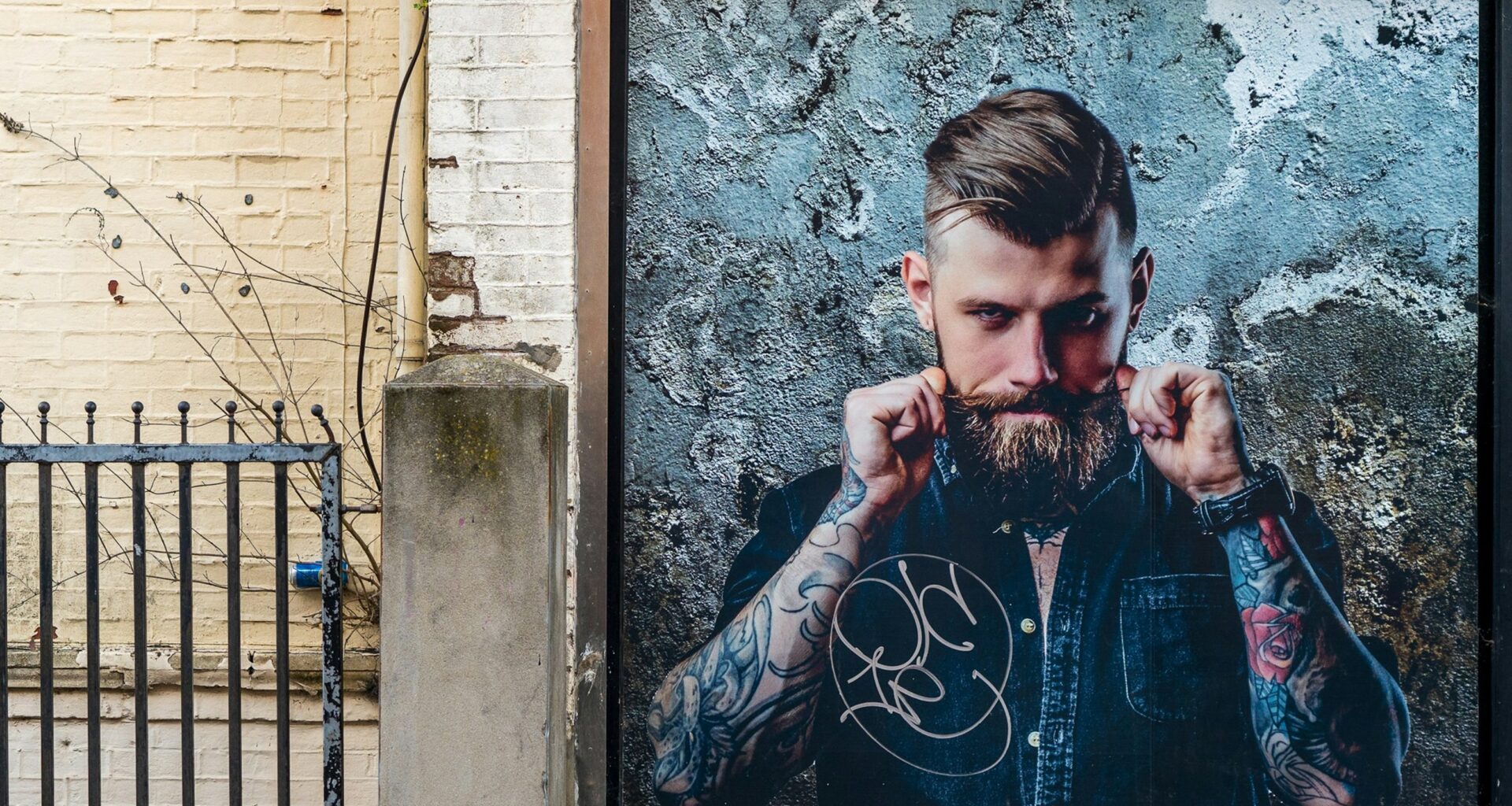 Homem tatuado com barba, posando contra parede cinza.