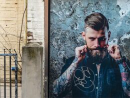Homem tatuado com barba, posando contra parede cinza.