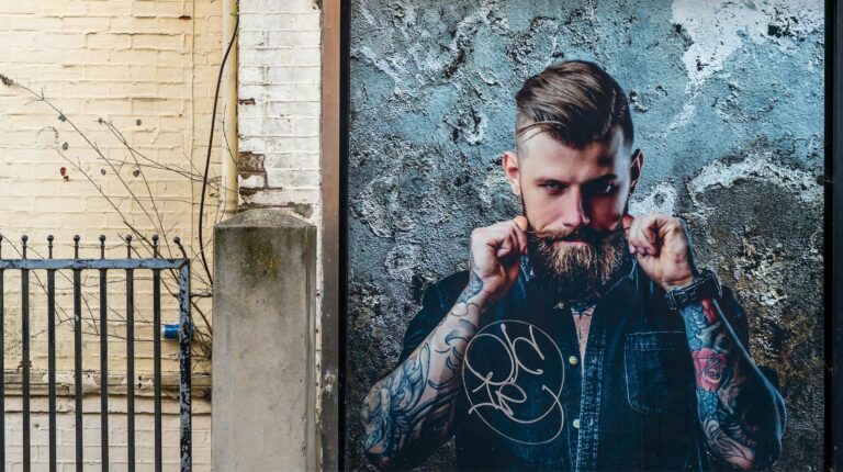 Homem tatuado com barba, posando contra parede cinza.