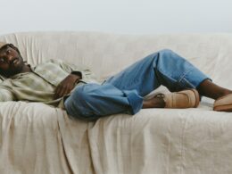 Homem jovem relaxando num sofá bege com roupa casual.