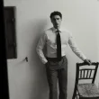 Homem de camisa e gravata, preto e branco, encostado a cadeira.