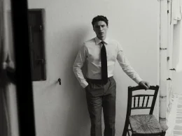 Homem de camisa e gravata, preto e branco, encostado a cadeira.
