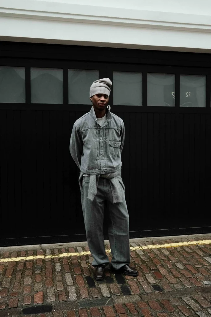 Homem com look ganga cinzenta e gorro, contra porta preta.