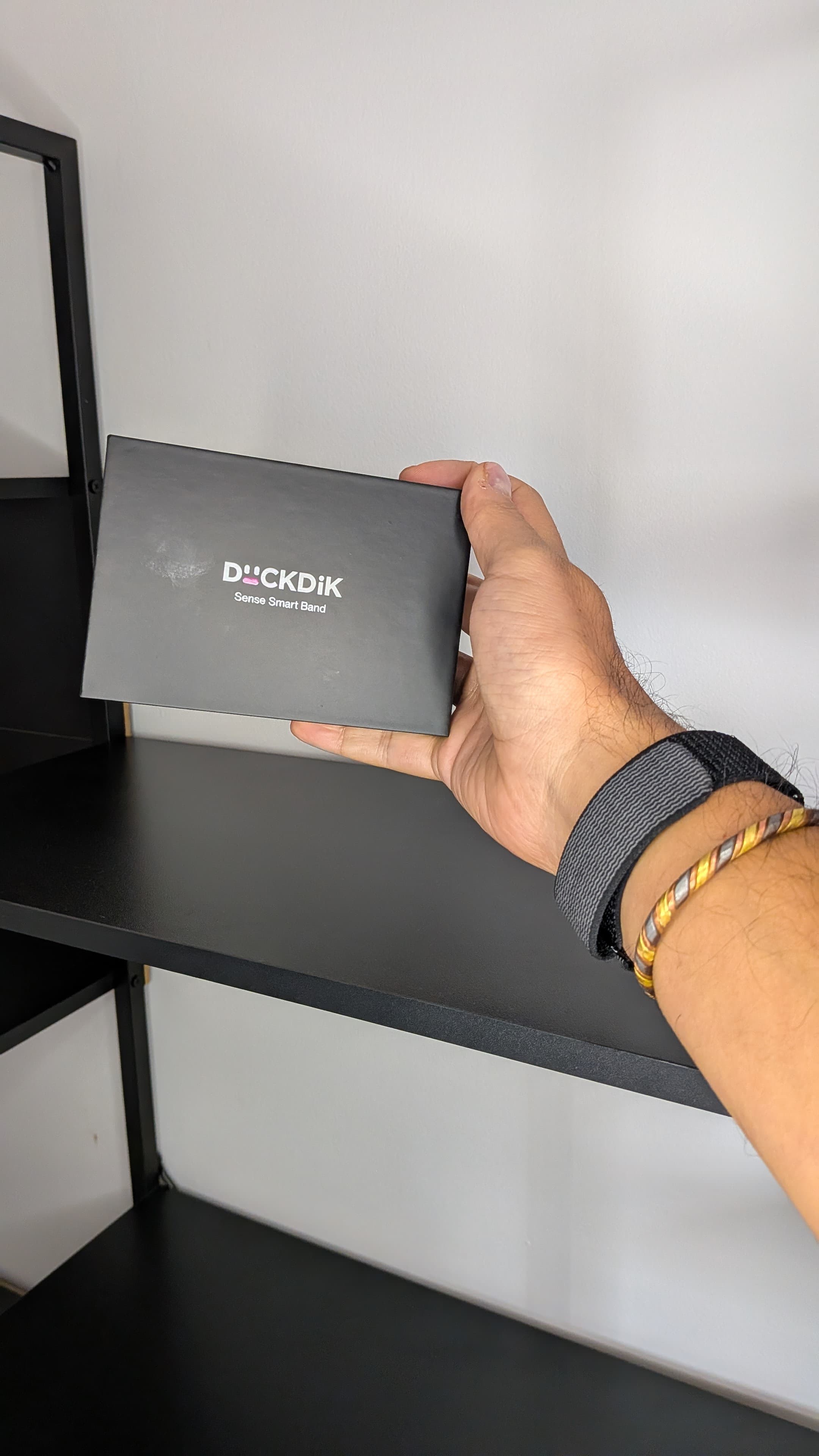 Mão com pulseiras a segurar a caixa preta da DuckDik Sense Smart Band junto a uma estante metálica