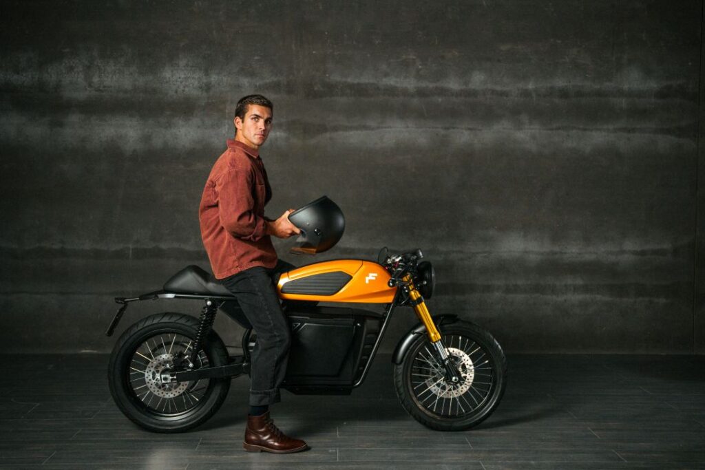 Homem com capacete ao lado de moto laranja.