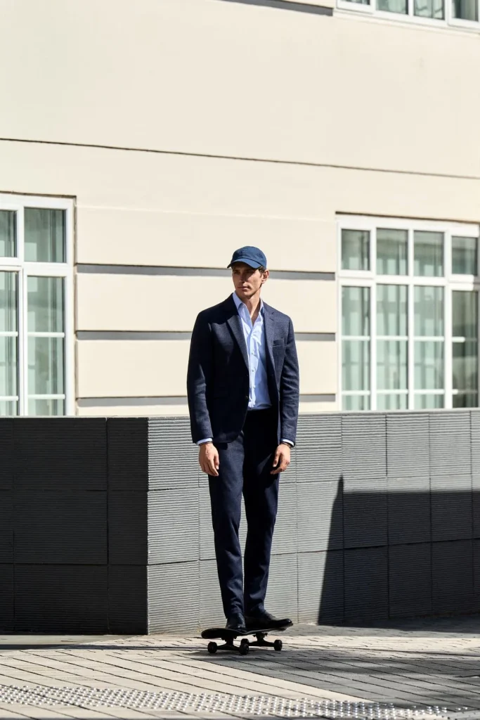 Homem de fato azul escuro, camisa clara e boné em cima de um skate num ambiente urbano, a vestir peças da coleção Hackett London outono inverno.