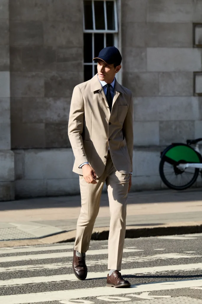 Homem a atravessar uma passadeira urbana a vestir um fato bege da coleção Hackett London outono inverno, combinado com camisa azul clara, gravata escura, boné azul e sapatos de pele castanhos.