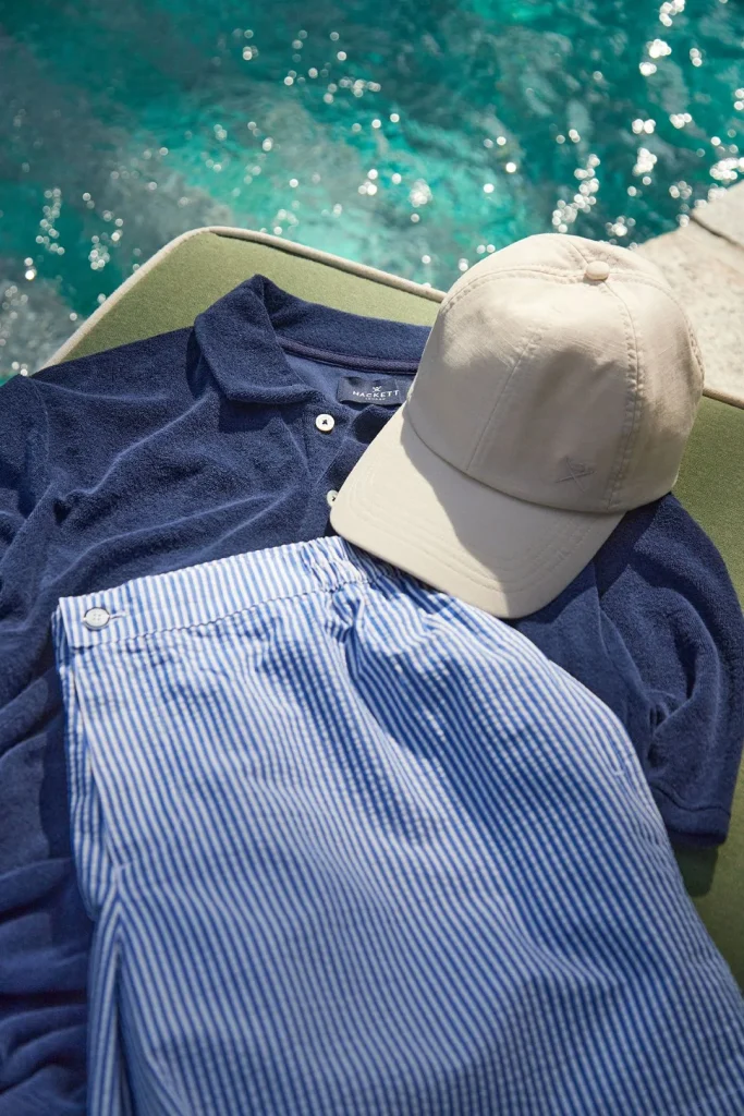 Polo azul, calções às riscas e boné junto à piscina.