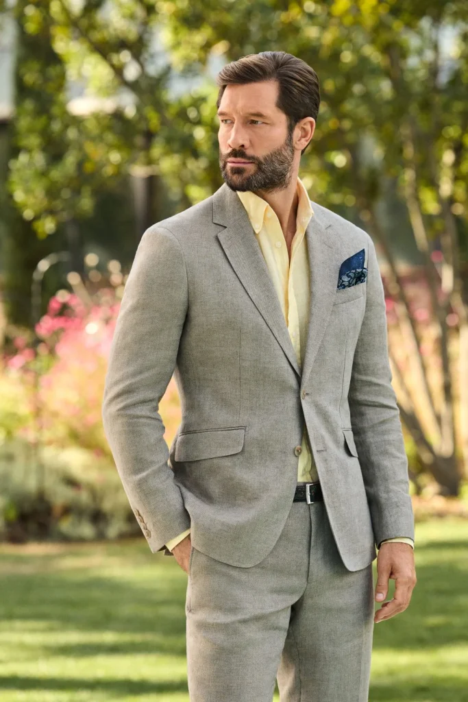 Homem com fato cinzento claro e camisa amarela num jardim.
