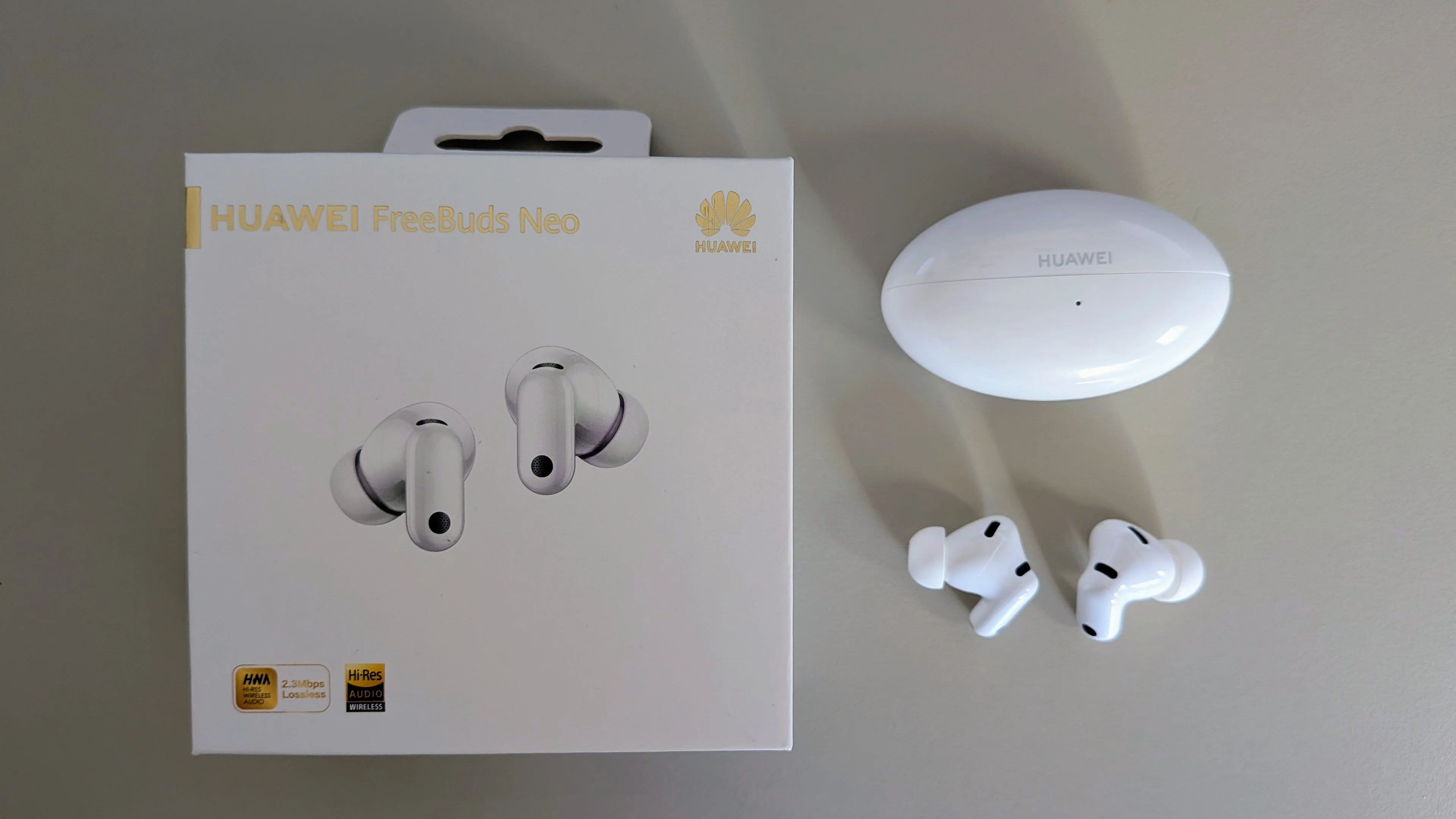 Caixa dos auriculares Huawei FreeBuds Neo ao lado do estojo de carregamento oval e dos auriculares brancos