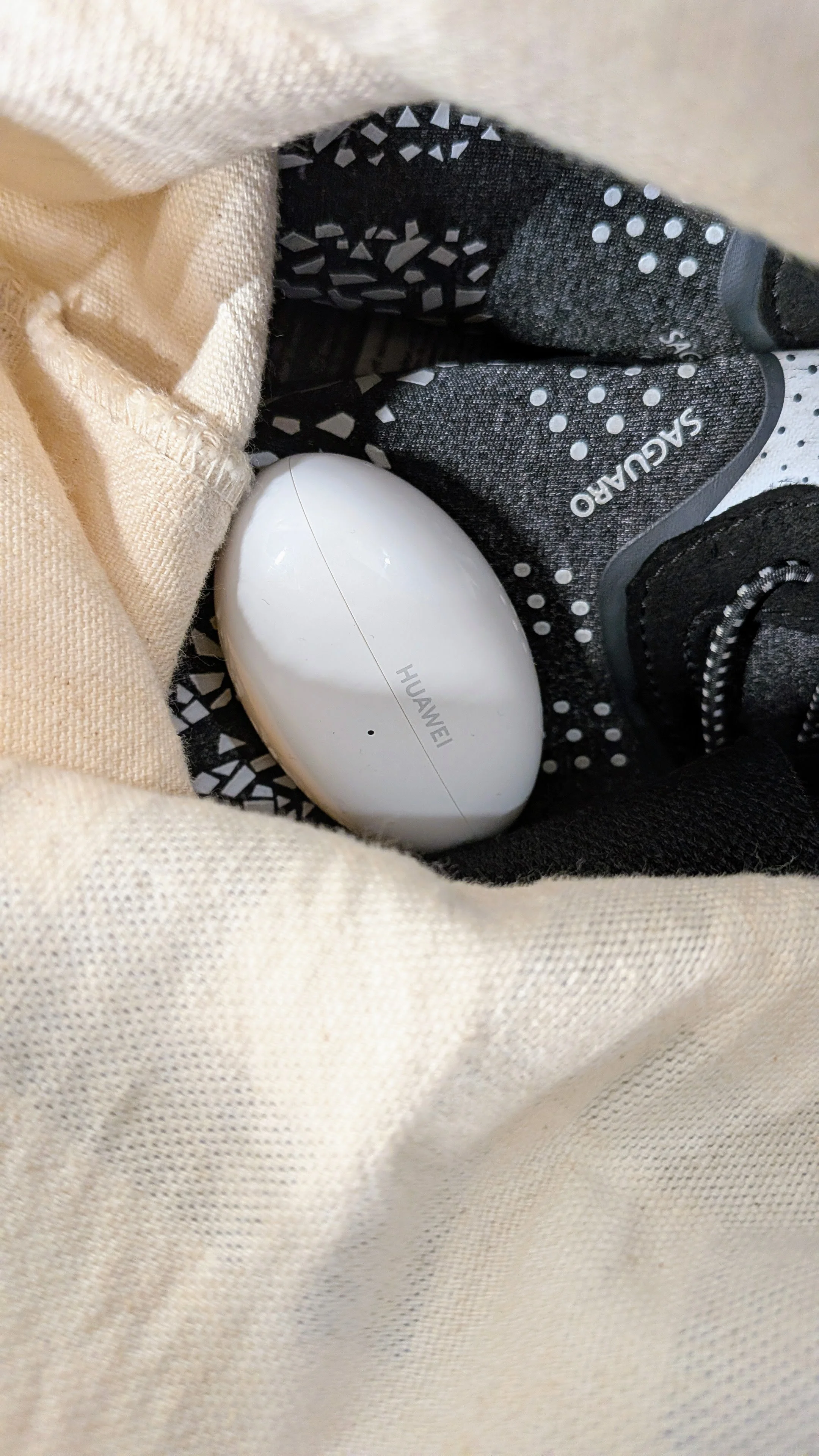Caixa de carregamento branca dos Huawei FreeBuds Neo dentro de um saco de tecido bege junto a uns ténis de desporto pretos.