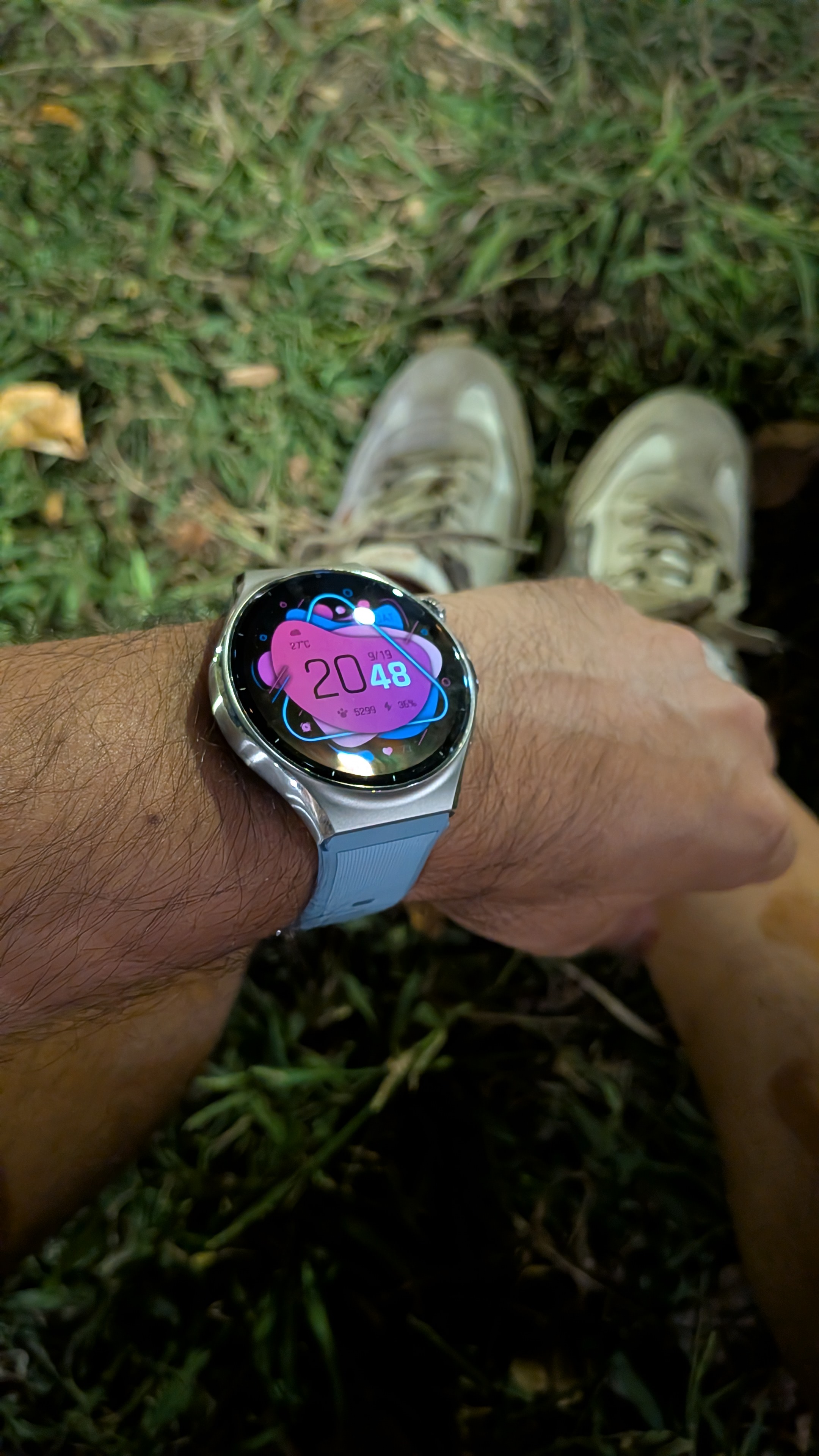 Braço de homem a usar o smartwatch Huawei Watch 6 com mostrador digital colorido e bracelete azul clara sobre fundo de relva.